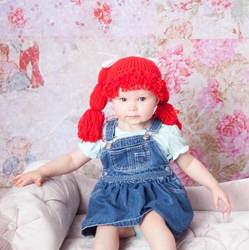 A toddler dressed in a Halloween costume, wearing a crocheted cabbage patch wig with yarn pigtails. The wig mimics the classic hairstyle of Cabbage Patch Kids dolls, giving the costume a cute and nostalgic feel."