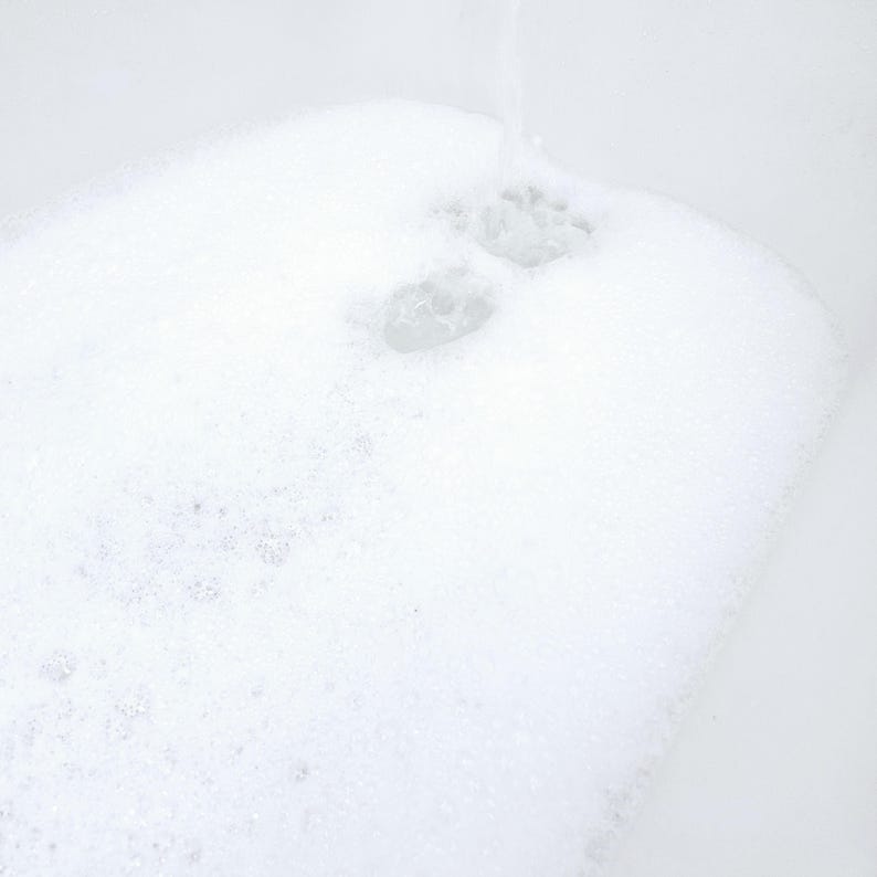 White bathtub filled with rich, fluffy foam created by NMJ Handmade Creations sulfate-free bubble bath. Thick layers of soft bubbles cover the water surface, showing the luxurious lather effect in a bright, clean setting.