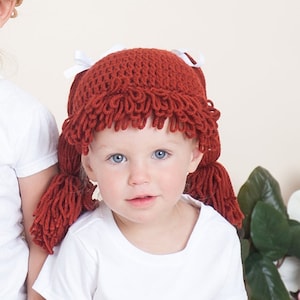 A baby dressed for Halloween is wearing a crocheted cabbage patch hat with yarn pigtails. The hat is styled to resemble the iconic Cabbage Patch Kids dolls, with curly yarn hair tied into two pigtails with bows.