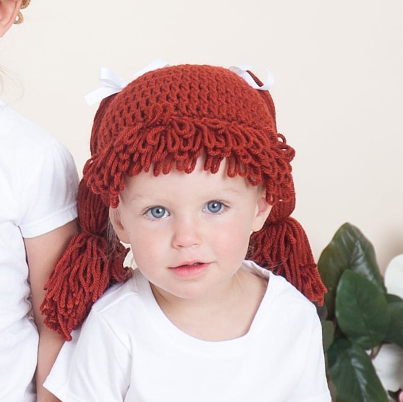 A baby dressed for Halloween is wearing a crocheted cabbage patch hat with yarn pigtails. The hat is styled to resemble the iconic Cabbage Patch Kids dolls, with curly yarn hair tied into two pigtails with bows.