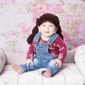 A baby wearing a handmade Halloween hat designed to look like a Cabbage Patch doll's hair. The crocheted hat mimics baby doll hair with yarn strands pulled neatly into pigtails, capturing the charming and playful essence of a Cabbage Patch Kids doll.