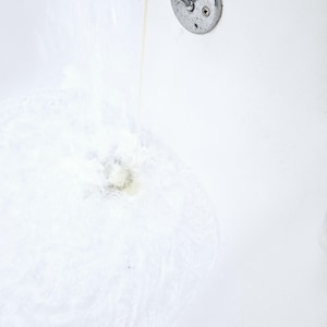 Close-up of clear water running into a white bathtub, activating NMJ Handmade Creations sulfate-free bubble bath. Gentle foaming action begins at the base, creating light bubbles and a refreshing, airy texture.