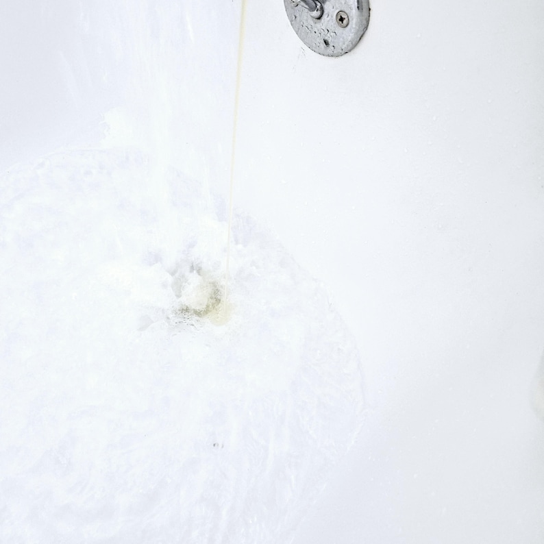 Close-up of clear water running into a white bathtub, activating NMJ Handmade Creations sulfate-free bubble bath. Gentle foaming action begins at the base, creating light bubbles and a refreshing, airy texture.