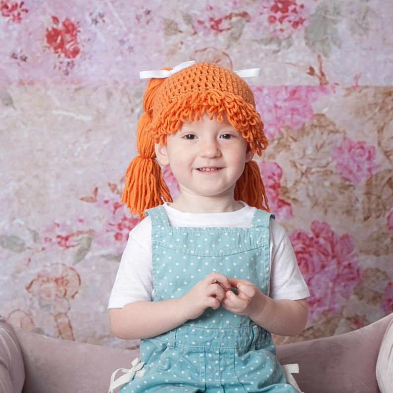 A baby wearing a handmade Halloween hat designed to look like baby doll hair from a Cabbage Patch Kid. The crocheted hat features textured yarn mimicking doll-like hair, giving the baby a charming, doll-inspired appearance