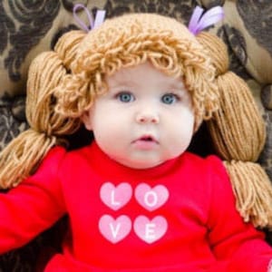 A toddler in a Halloween costume wearing a crocheted cabbage patch wig with yarn pigtails. The wig mimics the classic Cabbage Patch Kids doll hairstyle, with the pigtails tied off at the sides, giving the toddler a cute and nostalgic look.