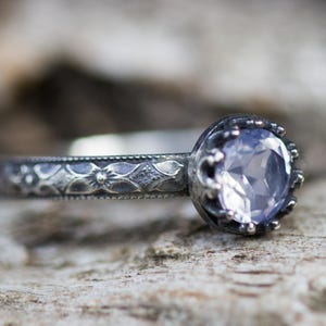 May include: A silver ring with a light purple gemstone, close up. The ring features an intricate floral pattern on the band and a crown-shaped setting around the central stone. The background is blurred and neutral.