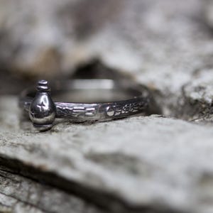 May include: A silver ring with a small bottle charm. The ring is engraved with a floral design.