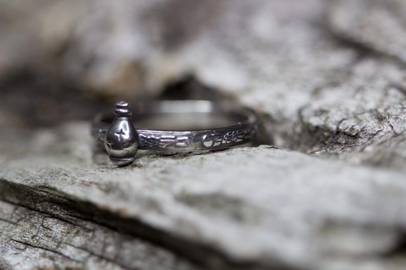 May include: A silver ring with a small bottle charm. The ring is engraved with a floral design.