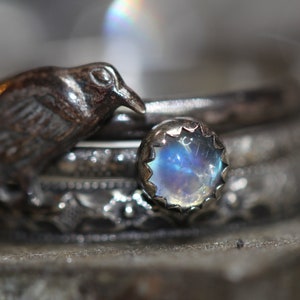 Puede incluir: Un anillo de plata con una piedra de luna y un peque&ntilde;o amuleto de p&aacute;jaro. El anillo est&aacute; apilado con otros anillos de plata.