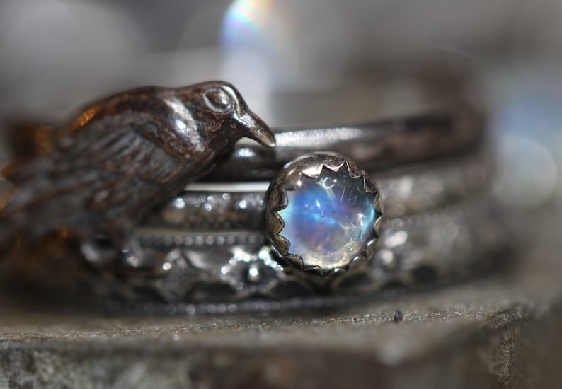 Puede incluir: Un anillo de plata con una piedra de luna y un peque&ntilde;o amuleto de p&aacute;jaro. El anillo est&aacute; apilado con otros anillos de plata.