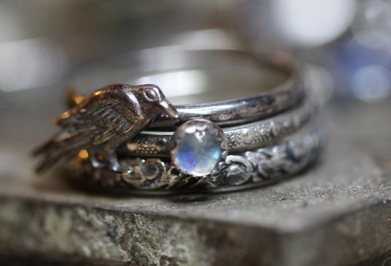 Puede incluir: Un anillo de plata con una piedra de luna y un peque&ntilde;o encanto de p&aacute;jaro. El anillo est&aacute; hecho de tres bandas, cada una con un dise&ntilde;o diferente.