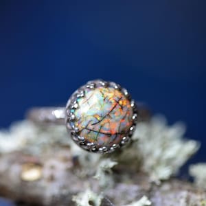 May include: A close-up of a silver ring with a large, round, colorful gemstone. The gemstone has a mix of orange, yellow, and green hues with dark veins. The ring is set on a natural, mossy surface against a blurred blue background.