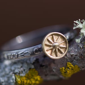 Puede incluir: Un anillo de plata con un diseño de sol dorado. El anillo presenta una banda detallada con patrones florales. El sol es un diseño circular con líneas radiantes, creando una apariencia de estrella. El anillo está sobre un fondo natural borroso.