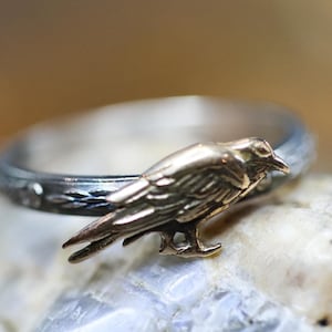 Peut inclure: Bague en argent ornée d'un corbeau doré. L'anneau présente une finition texturée gris foncé. Le corbeau, aux détails complexes, se détache de l'argent, créant un contraste saisissant.