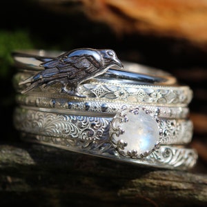 May include: A set of five silver rings with intricate floral designs. The top ring features a silver raven perched on a band. The bottom ring features a large, iridescent moonstone.