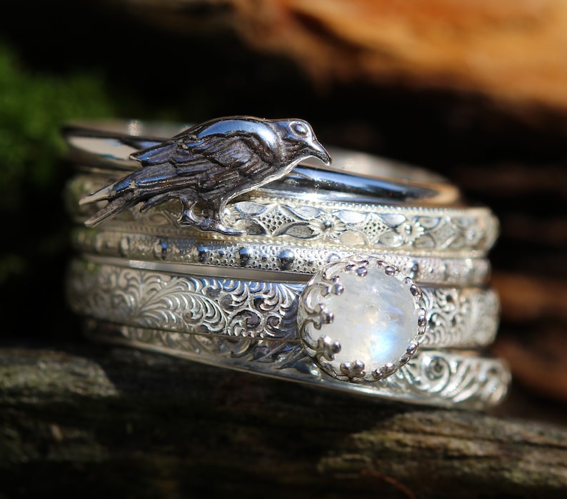 May include: A set of five silver rings with intricate floral designs. The top ring features a silver raven perched on a band. The bottom ring features a large, iridescent moonstone.
