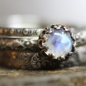 May include: A silver ring with a moonstone set in a delicate, ornate crown design. The ring is stacked with three other silver bands, each with a different textured pattern.
