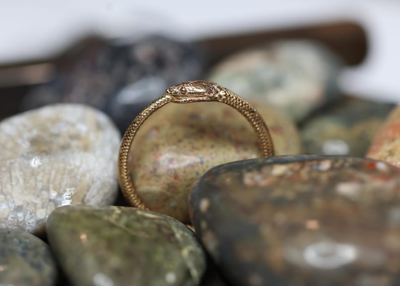 May include: A gold-colored ring in the shape of a snake, with the head of the snake at the top of the ring. The ring is resting on a collection of smooth, colorful stones. The ring has a textured pattern.