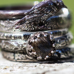 Peut inclure: Une pile d'anneaux en argent avec des motifs complexes, y compris un anneau avec une pierre noire et un anneau avec un motif d'oiseau.