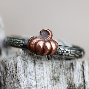 May include: A silver ring featuring a small, three-dimensional copper pumpkin centerpiece. The band of the ring is oxidized silver and displays a detailed, embossed floral pattern that wraps around the circumference of the metal band.