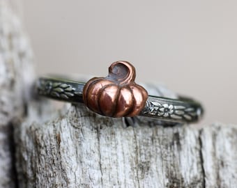 May include: A silver ring featuring a small, three-dimensional copper pumpkin centerpiece. The band of the ring is oxidized silver and displays a detailed, embossed floral pattern that wraps around the circumference of the metal band.