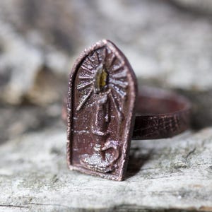 Puede incluir: Un anillo de metal marrón con un diseño detallado de una ventana con una vela en su interior. El anillo está sobre una superficie de madera.