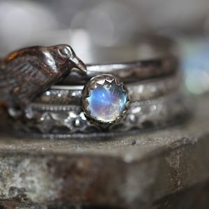 Puede incluir: Un anillo de plata con una piedra de luna, con un dise&ntilde;o de cuervo detallado. El anillo est&aacute; apilado con otras bandas de plata.