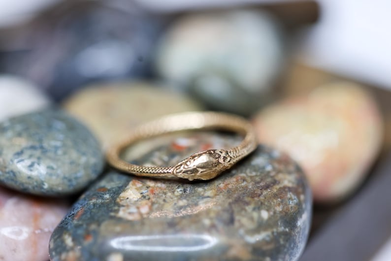 May include: A gold-colored ring in the shape of a snake, with detailed scales and a textured head. The ring is resting on a collection of smooth, colorful stones, creating a natural and elegant display.