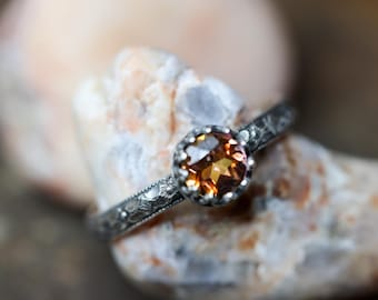 May include: A silver ring with an orange gemstone. The ring features a detailed band with a textured pattern. The gemstone is round and set in a decorative bezel. The ring is displayed on a light-colored stone.