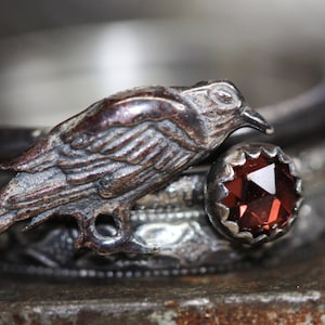 Peut inclure: Une bague en argent avec un motif d'oiseau détaillé et une pierre précieuse rouge. La bague est sertie sur une bande en argent texturée.