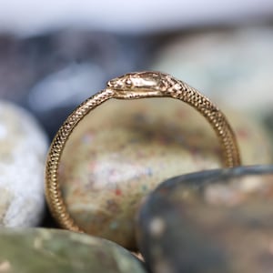 May include: A gold-colored ring in the shape of a snake, with a textured surface. The ring is resting on a bed of smooth, colorful stones. The snake's head is detailed and facing forward.
