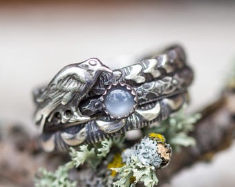 May include: A stack of silver rings with intricate dragon and bird designs. The top ring features a bird's head, while the center ring holds a round, pale blue gemstone. The rings are set against a blurred natural background.