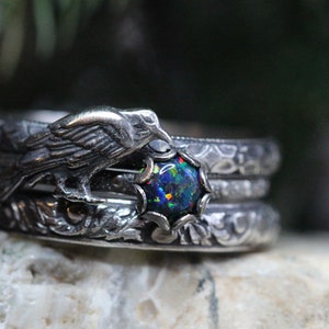 Puede incluir: Un anillo de plata con un cuervo posado sobre una piedra de &oacute;palo redonda e iridiscente. El anillo est&aacute; intrincadamente detallado con patrones florales.