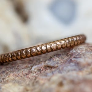 May include: A hammered copper ring with a textured surface. The ring is a thin band, likely a wedding band or stacking ring. The ring is displayed on a rough, multi-colored stone.