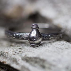 Peut inclure: Une bague en argent avec un motif détaillé d'une petite bouteille avec un bouchon. La bague est posée sur une surface en bois.