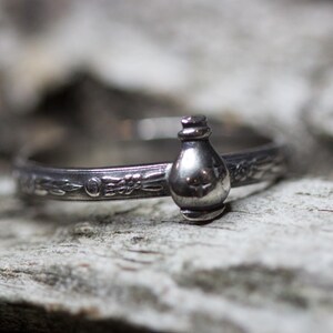 May include: A silver ring with a detailed design featuring a small, round bottle charm.