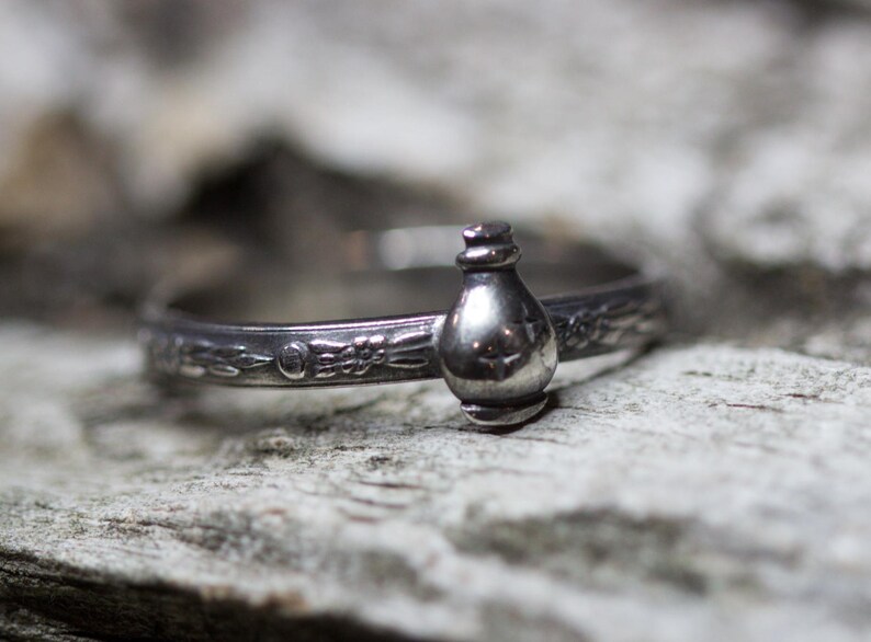 May include: A silver ring with a detailed design featuring a small, round bottle charm.