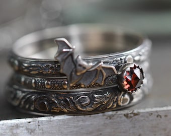 May include: A stack of three silver rings. The top ring features a bat design, and the second ring has a red gemstone. The rings have intricate floral and dot patterns, creating a gothic or fantasy aesthetic.