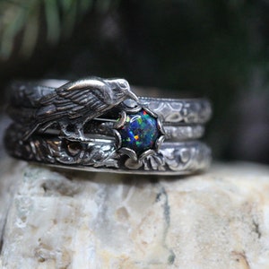 Puede incluir: Un anillo de plata con un cuervo posado en una rama, sosteniendo una piedra preciosa de &oacute;palo negro. El anillo est&aacute; intrincadamente detallado con patrones florales.