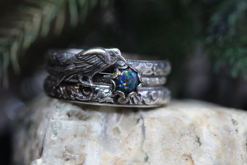 Puede incluir: Un anillo de plata con un cuervo posado en una rama, sosteniendo una piedra preciosa de &oacute;palo negro. El anillo est&aacute; intrincadamente detallado con patrones florales.