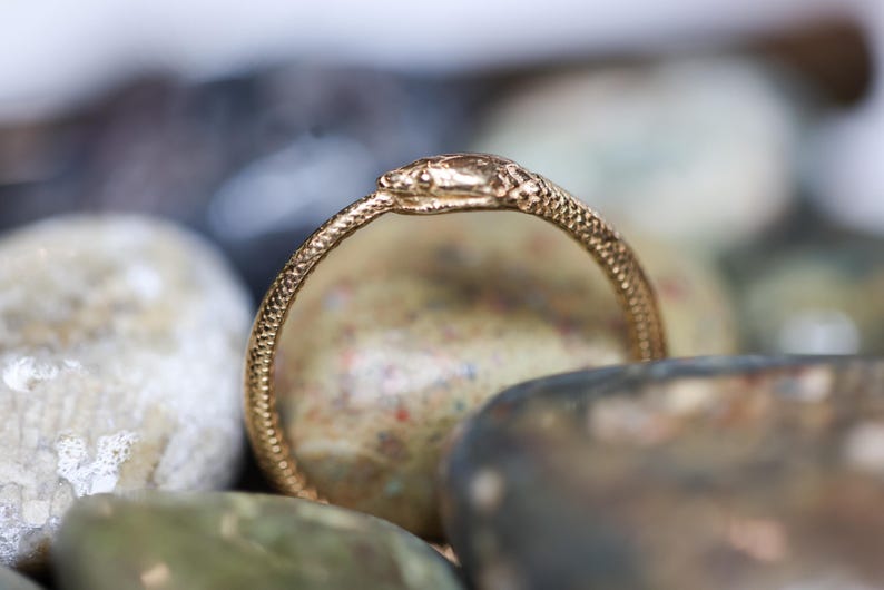 May include: A gold-colored ring in the shape of a snake, with a textured surface. The ring is resting on a bed of smooth, colorful stones. The snake's head is detailed and facing forward.
