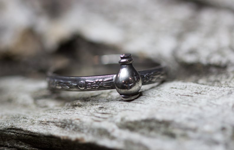 May include: A silver ring with a detailed design featuring a small bottle charm.