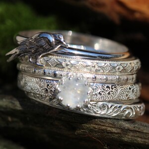 May include: A set of five silver rings with intricate engraved designs. The top ring features a small bird perched on the band. The ring set is on a brown wooden surface.