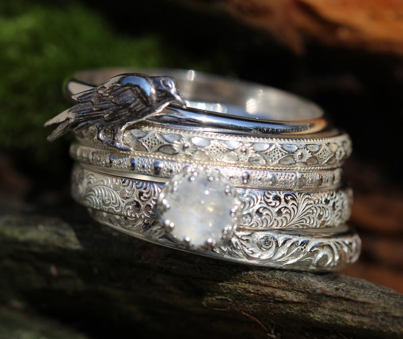 May include: A set of five silver rings with intricate engraved designs. The top ring features a small bird perched on the band. The ring set is on a brown wooden surface.