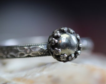 May include: A silver ring with a round, textured center piece and a detailed band.