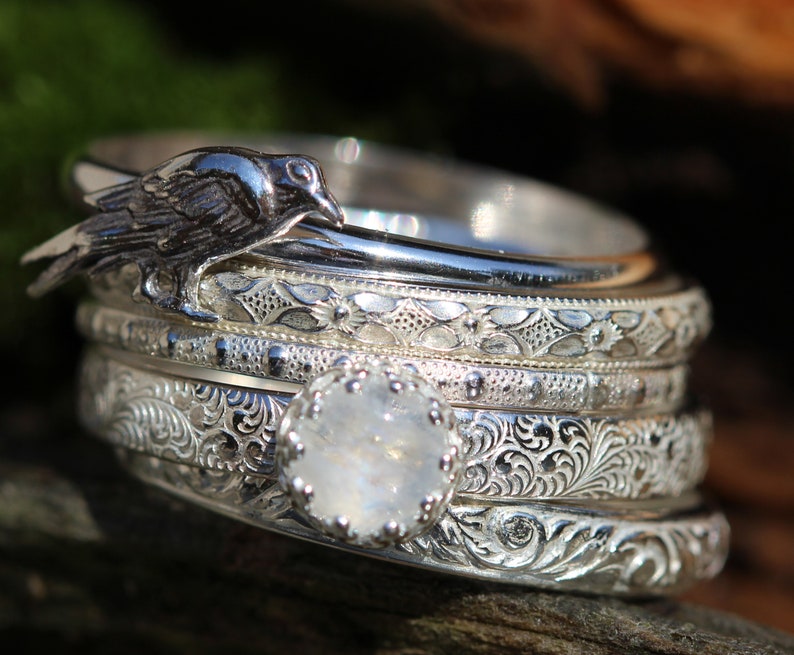 May include: A set of five silver rings with intricate engraved designs. The top ring features a silver crow perched on the band. The bottom ring features a large, round, white gemstone.
