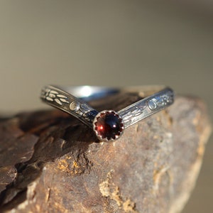 May include: A silver ring with a V-shaped band and a red gemstone in the center. The band is decorated with a floral design.
