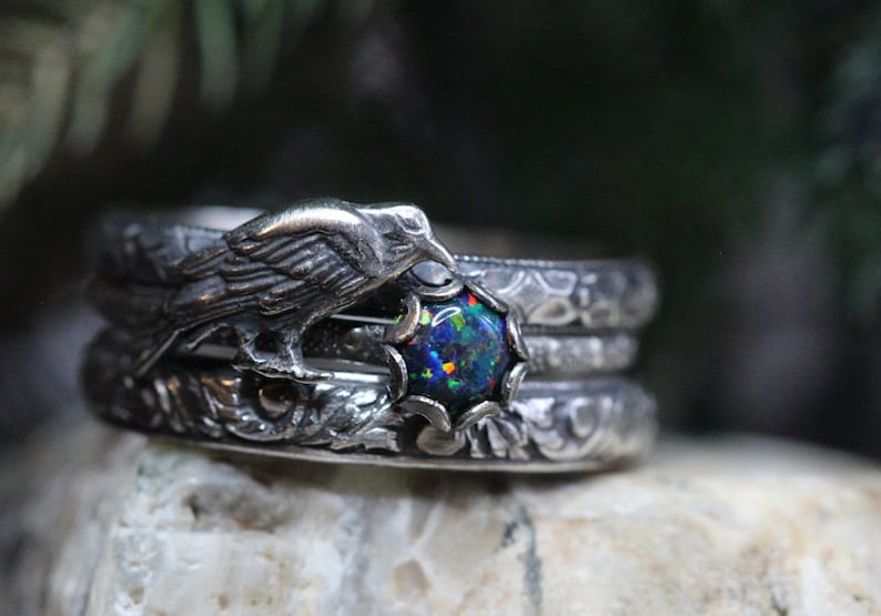 Puede incluir: Un anillo de plata con un cuervo posado sobre una piedra de &oacute;palo redonda e iridiscente. El anillo est&aacute; intrincadamente detallado con patrones florales.