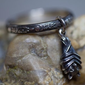 May include: A silver ring featuring a detailed floral design and a dangling bat charm. The bat is hanging upside down with its wings folded.