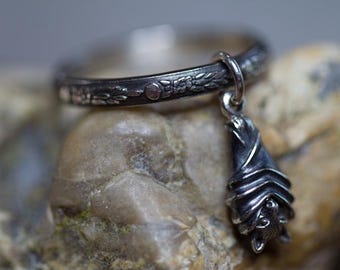 May include: A silver ring with a detailed floral design and a dangling bat charm. The bat is hanging upside down with its wings folded.